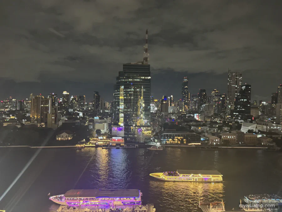 Illuminated passenger boats on the Chao Phraya with buildings on the opposite bank