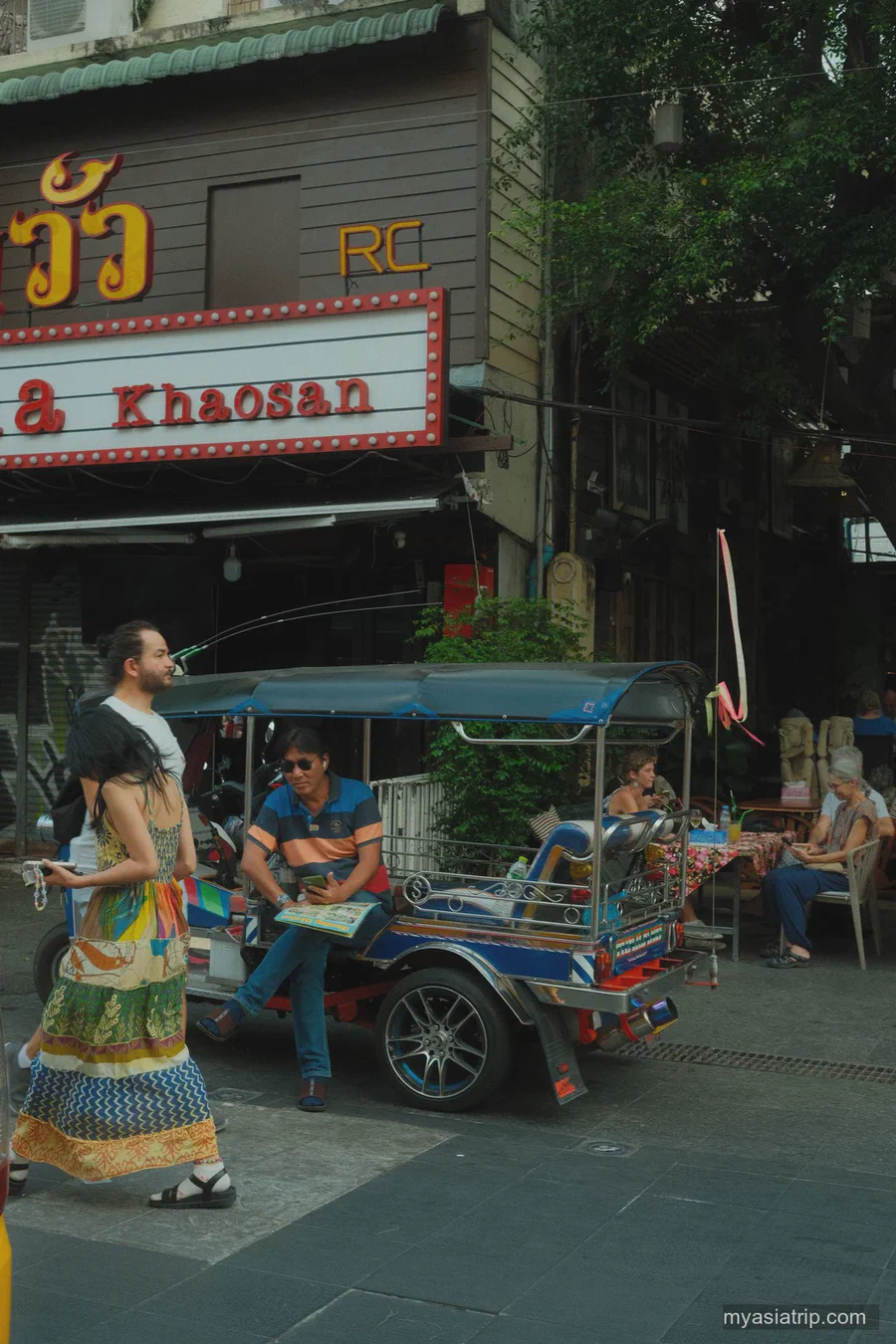 A tuk-tuk and pedestrians outside a Khaosan storefront in daylight